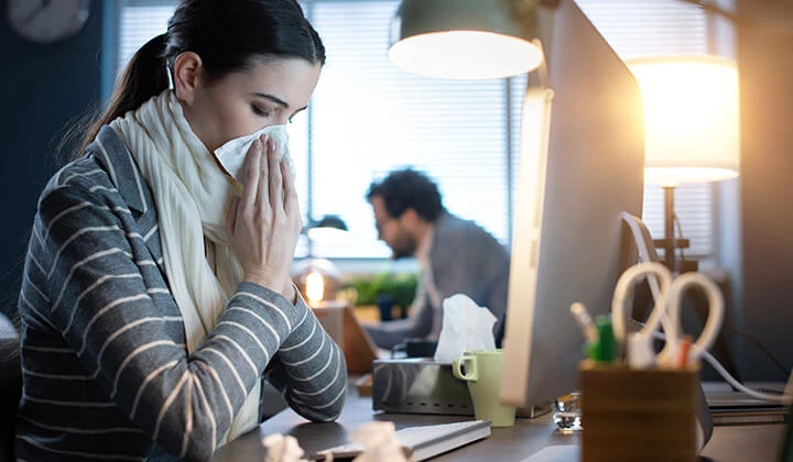 Frau trotz Erkältung auf der Arbeit Verschleppte Erkältung auskurieren und arbeiten vermeiden