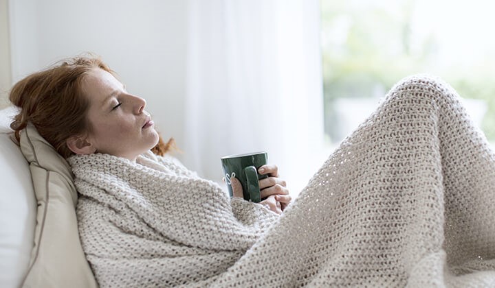 Frau mit Tee unter einer Decke Verschiedene Maßnahmen eignen sich, um eine Erkältung vorbeugen zu können